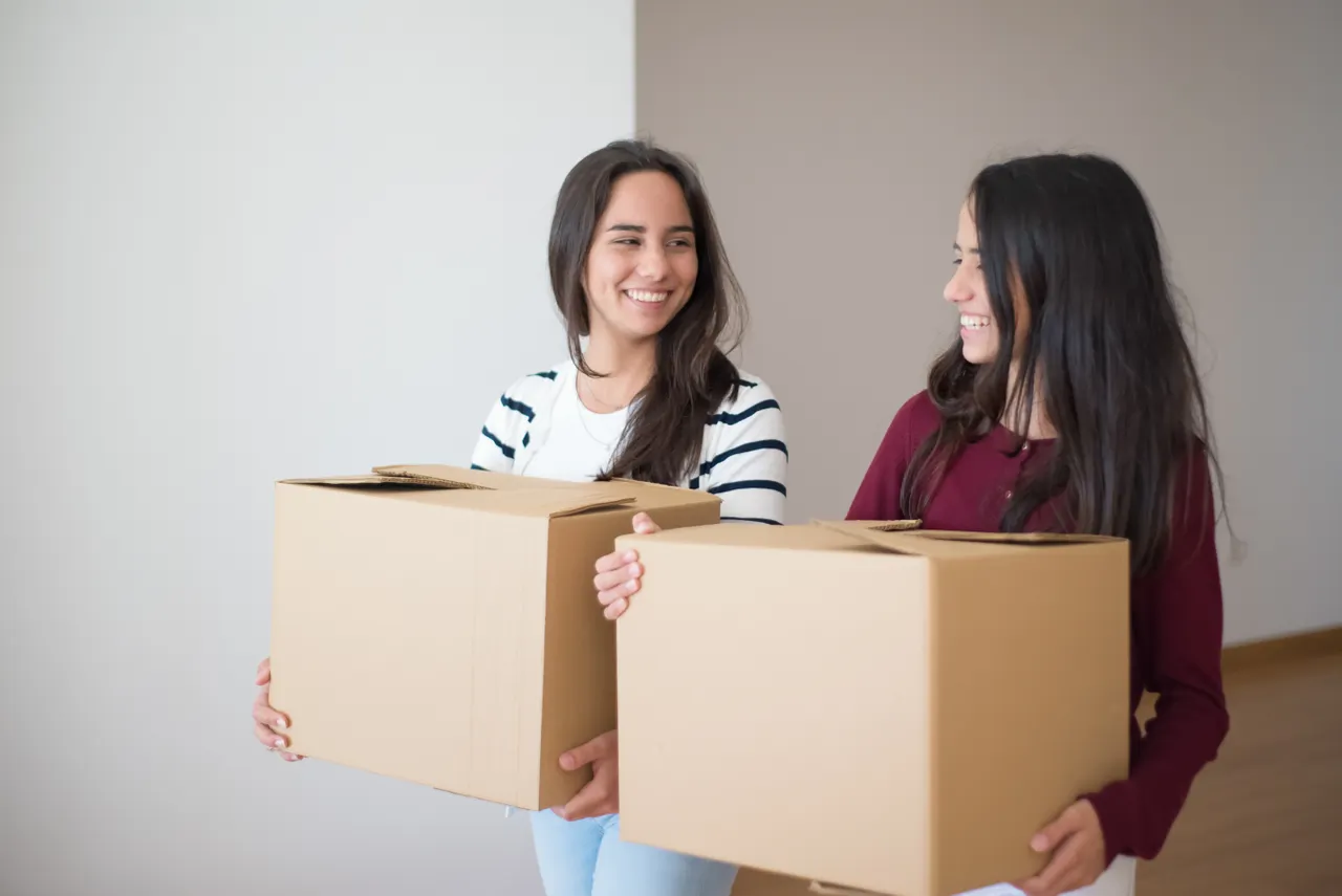 Two women carrying plain cardboard boxes inside a home